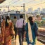 Editorial illustration of workers on an Indian suburban railway platform, with homes and office buildings beyond.