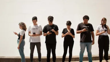 Six young people standing in a row against a wall, every one of them looking at a phone