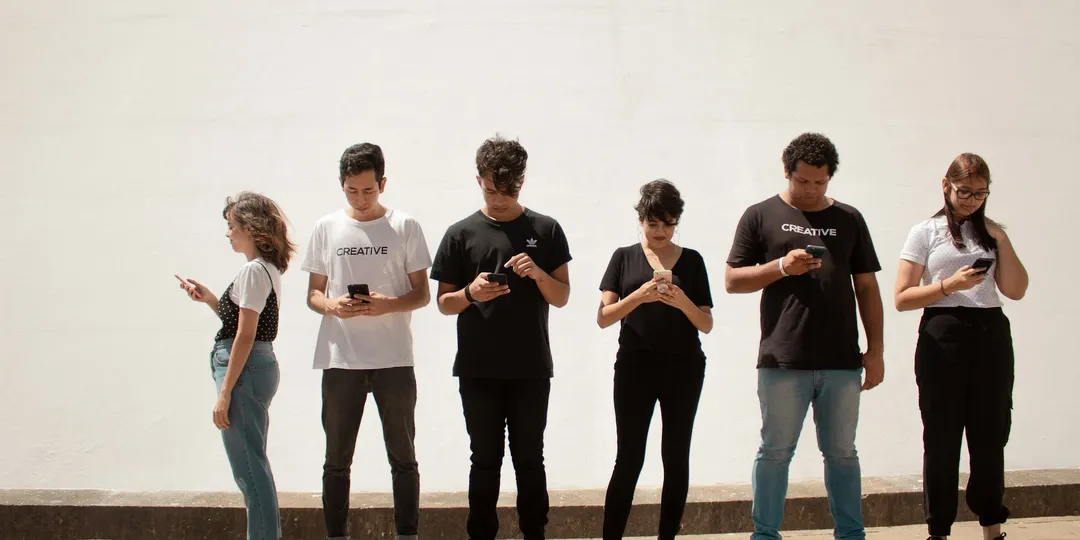 Six young people standing in a row against a wall, every one of them looking at a phone