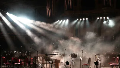 An empty stage set with microphones and music stands, spotlights cutting through haze