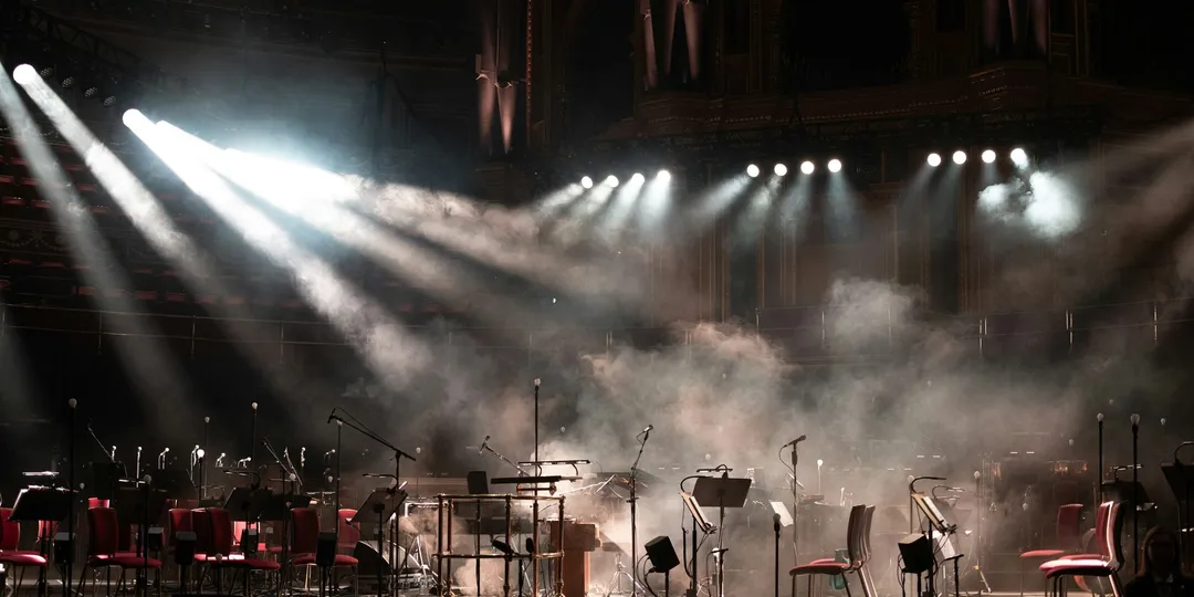 An empty stage set with microphones and music stands, spotlights cutting through haze