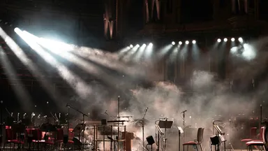 An empty stage set with microphones and music stands, spotlights cutting through haze
