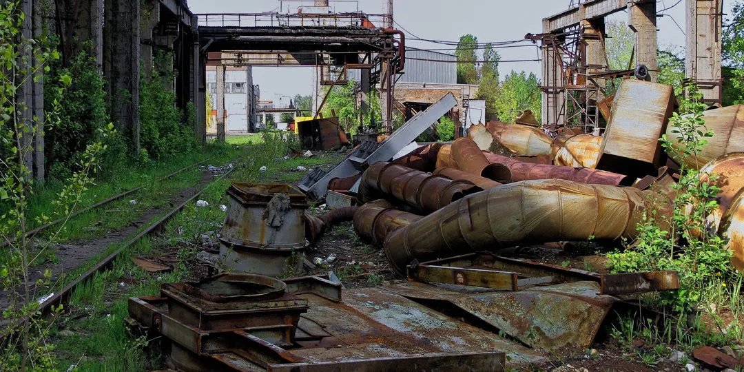A derelict factory yard with rusted machinery and overgrown ground