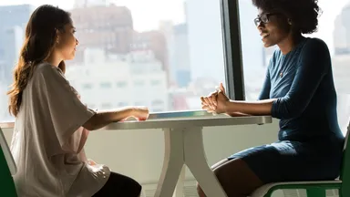 Two people sitting across a table in conversation beside a window