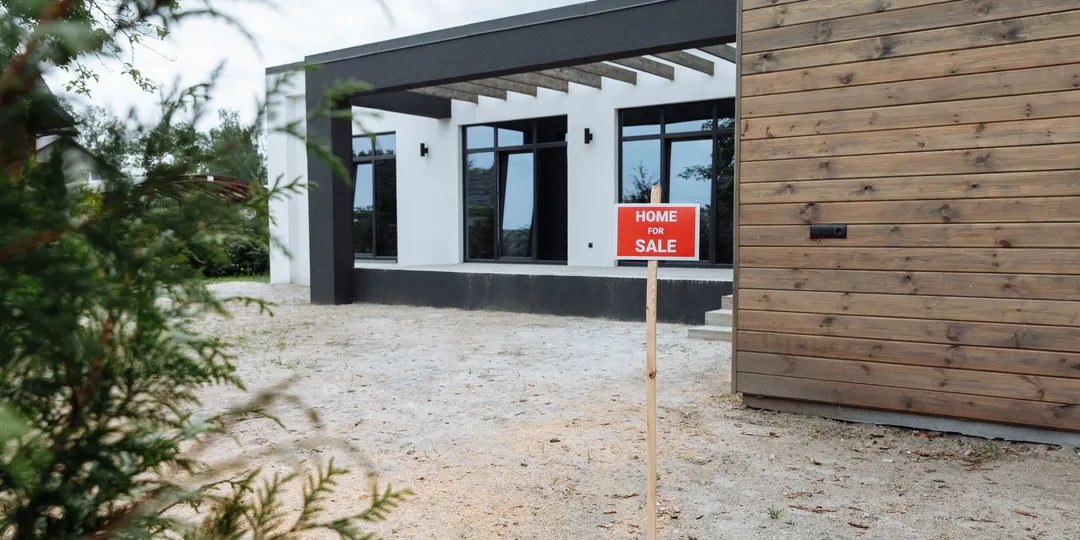 A red 'Home For Sale' sign staked in the gravel yard of a newly built house