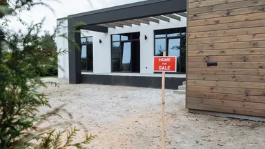 A red 'Home For Sale' sign staked in the gravel yard of a newly built house
