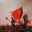 A dense crowd waving flags amid red smoke from flares